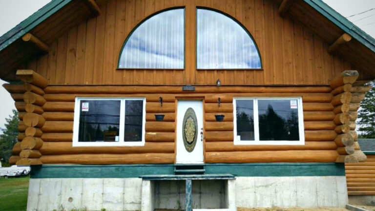 Chalet en bois rond avec grande fenêtre cintrée et porte blanche décorative, situé sur des fondations en béton.