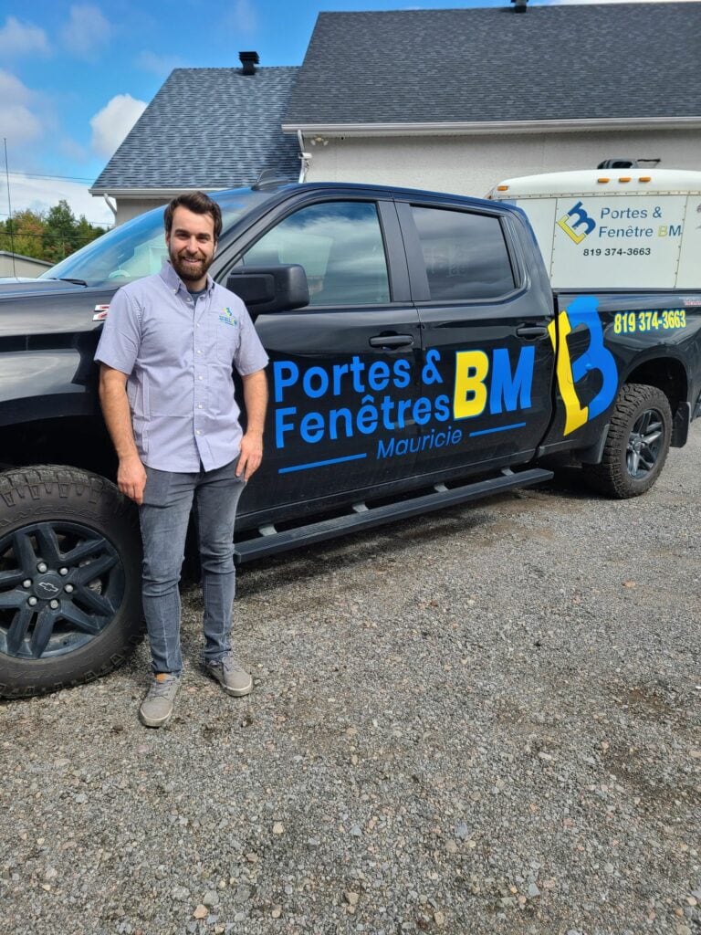 Un homme souriant devant un camion de Portes & Fenêtres BM à l'extérieur, ciel bleu et bâtiment en arrière-plan.