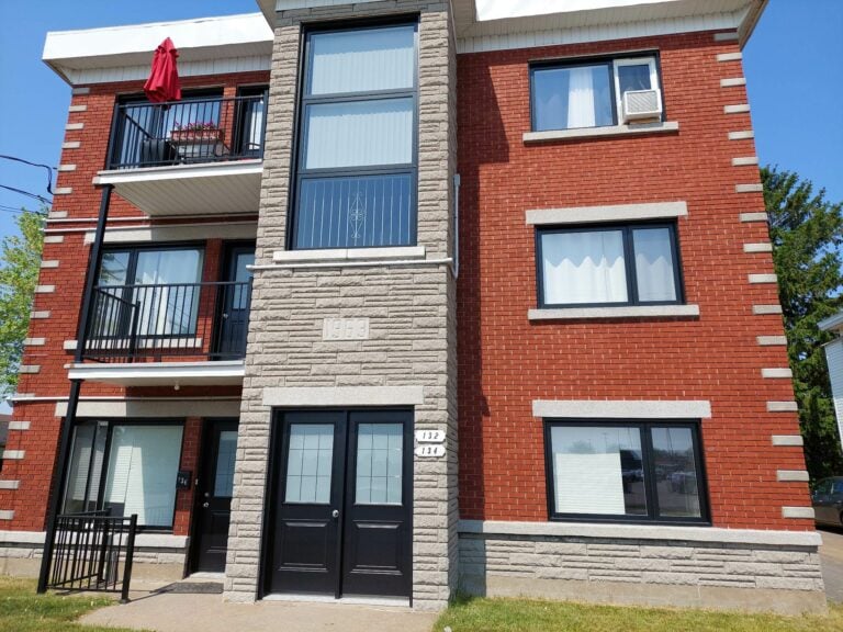 Immeuble résidentiel en briques rouges avec balcons et grandes fenêtres, vu sous un ciel bleu clair.