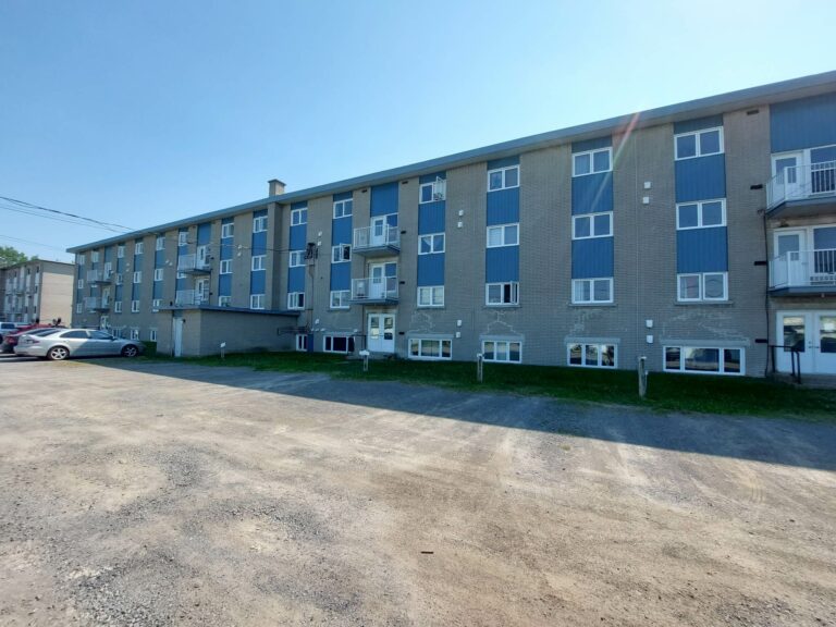 Immeuble résidentiel en brique avec balcons, parking et voitures sous un ciel bleu clair.