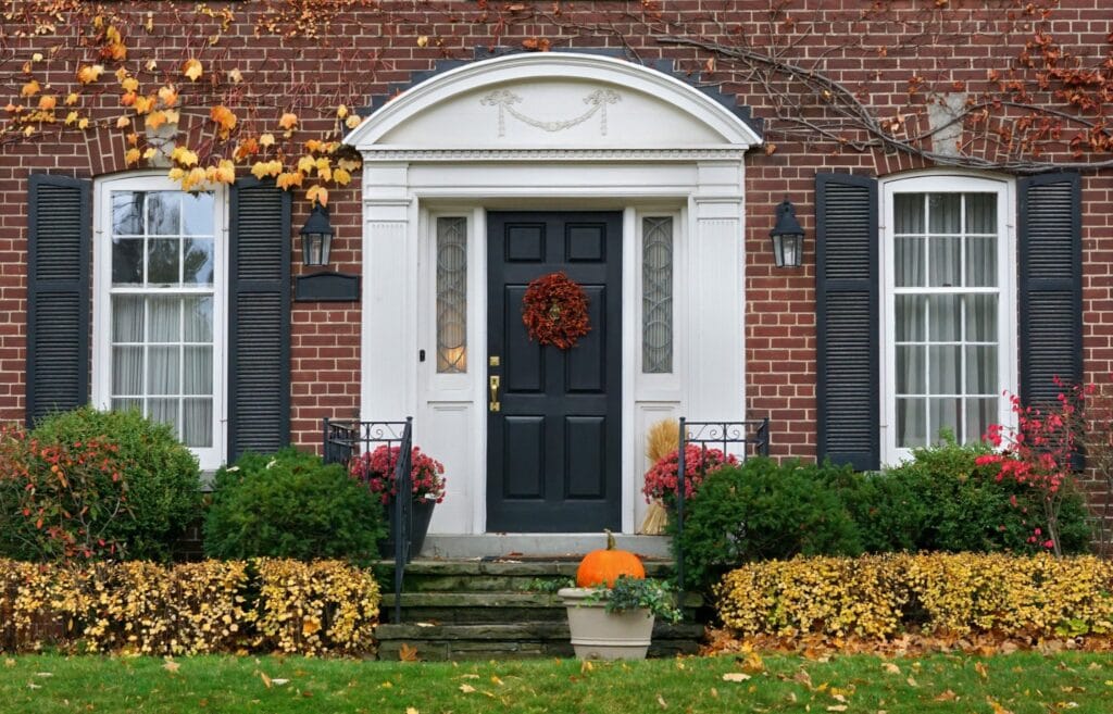 Porte et fenetre maison en brique avec porte noire ornée d'une couronne de Noël, entourée de fenêtres à volets noirs, décoration automnale, porte d'entrée élégante, service de pose portes et fenêtres, ambiance chaleureuse et festive.