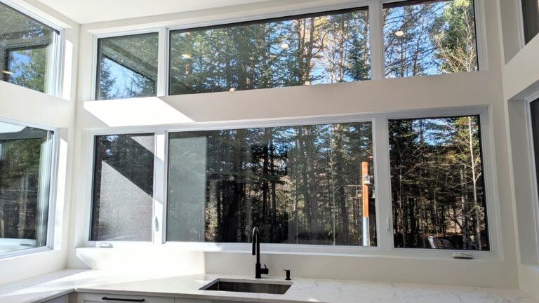 Vue intérieure d'une grande baie vitrée avec cadre blanc, offrant une vue dégagée sur la forêt extérieure, idéale pour renforcer la luminosité et l'isolation thermique.